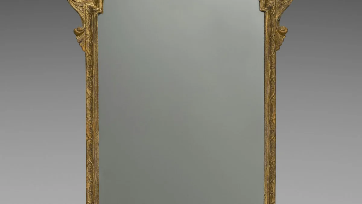 Carved gilt crest and moldings of an ornate antique gilded mirror against a plain grey wall. — a The Obsidian Key guide to Ask Who Ran It Before You: Lineage as Provenance.